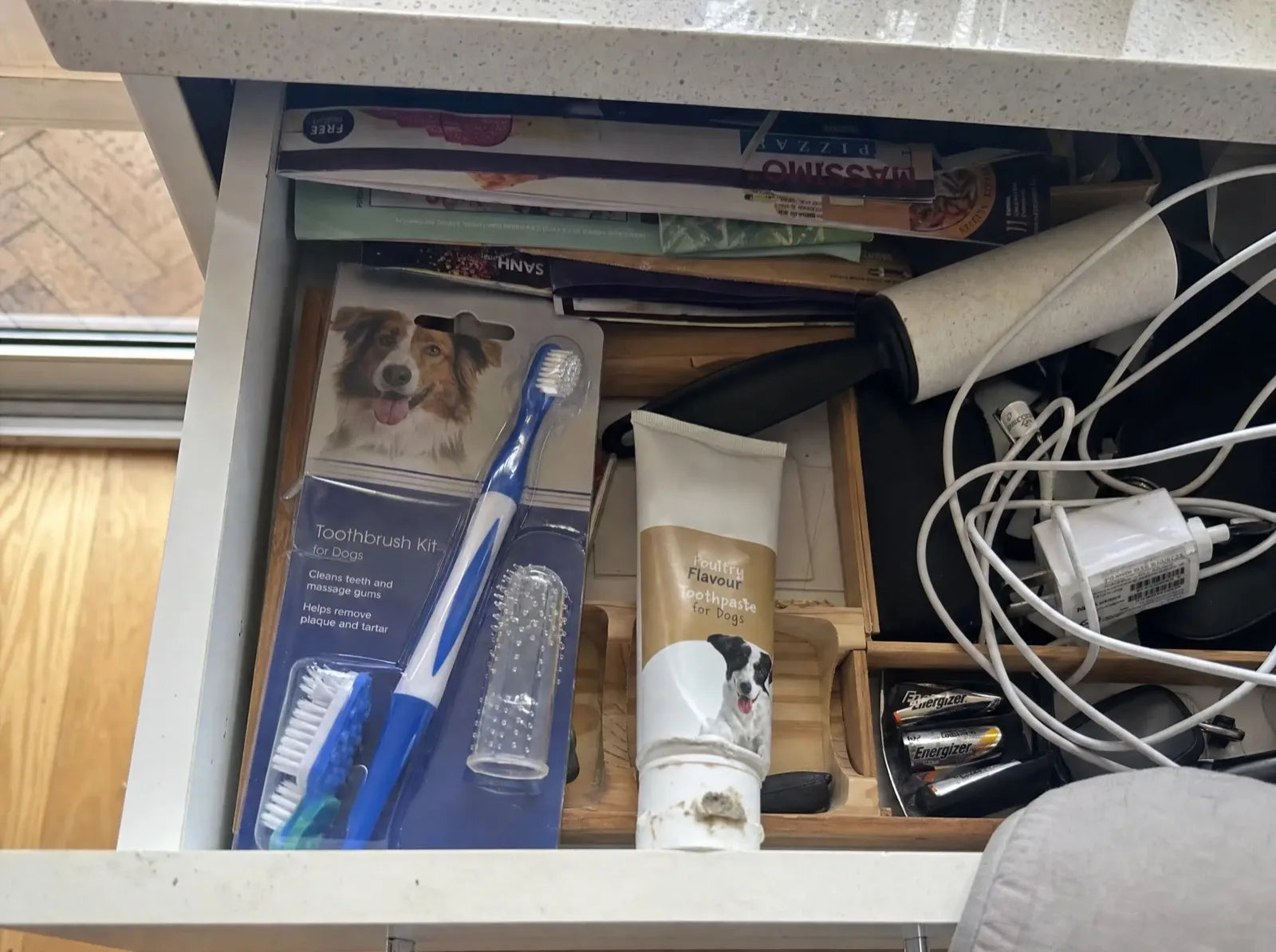 A half-open kitchen drawer with an unused dog toothbrush kit pushed behind takeaway menus and cables